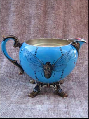 A whimsical blue tea set creamer with a curved decorative handle, adorned with a large golden cicada and hand painted details