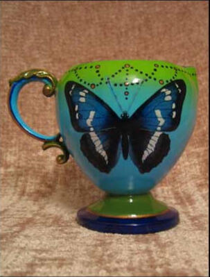 A whimsical aqua fading to green tea set creamer with a curved decorative handle, adorned with a large blue butterfly and hand painted details (front view)