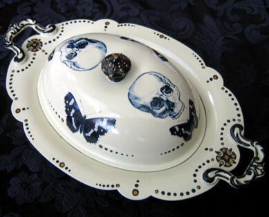 A whimsical cream colored antique butter dish with curved ornate handles, and gold accents adorned with skulls, butterflies, and hand painted details.