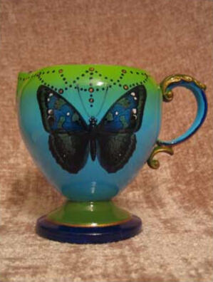A whimsical aqua fading to green tea set creamer with a curved decorative handle, adorned with a large blue butterfly and hand painted details (back view)
