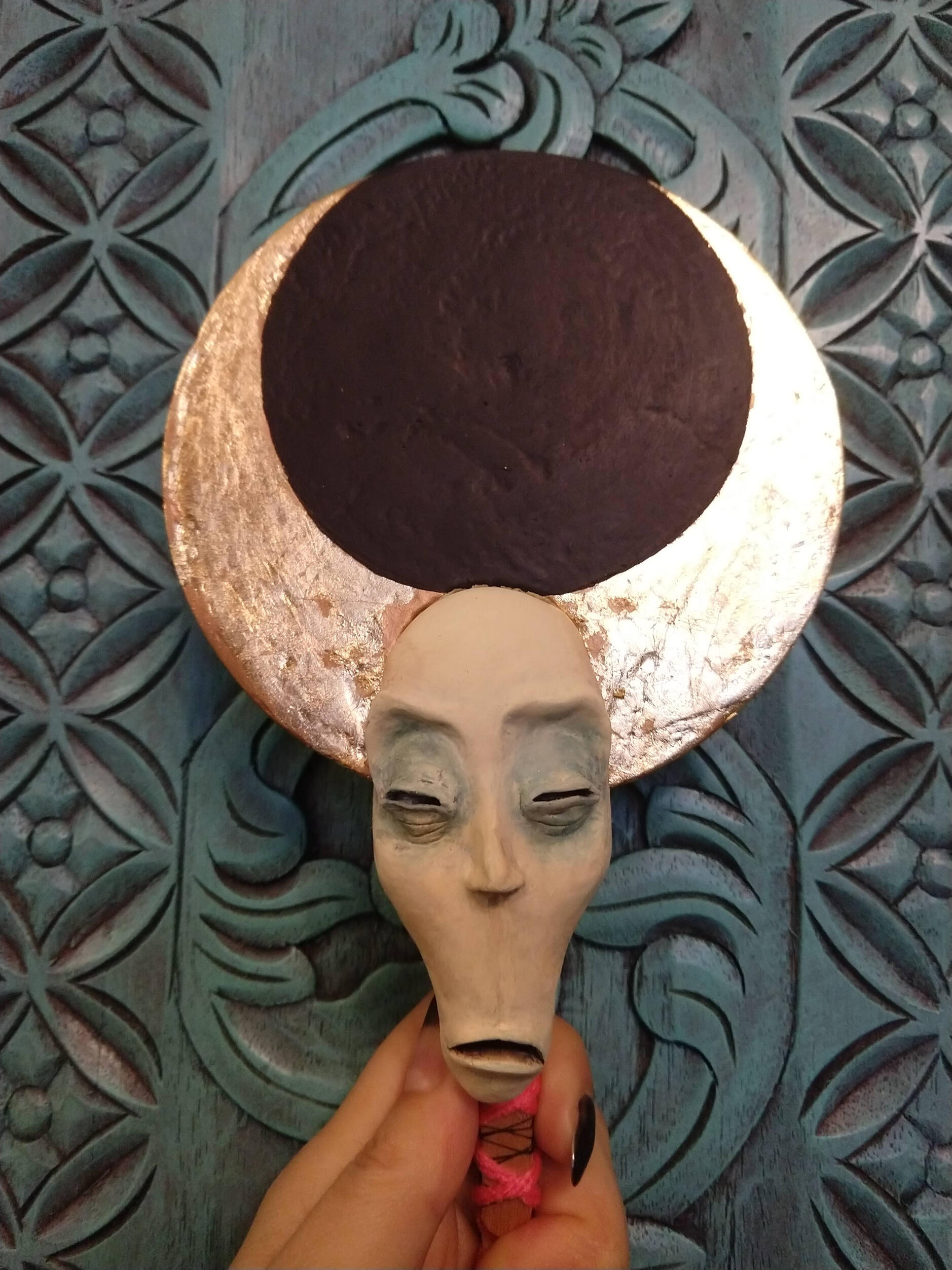 A paper mache doll, with a long pale face, squinting judgmentally in your direction. A large gold-leaf embellished crescent moon disk atop it's head