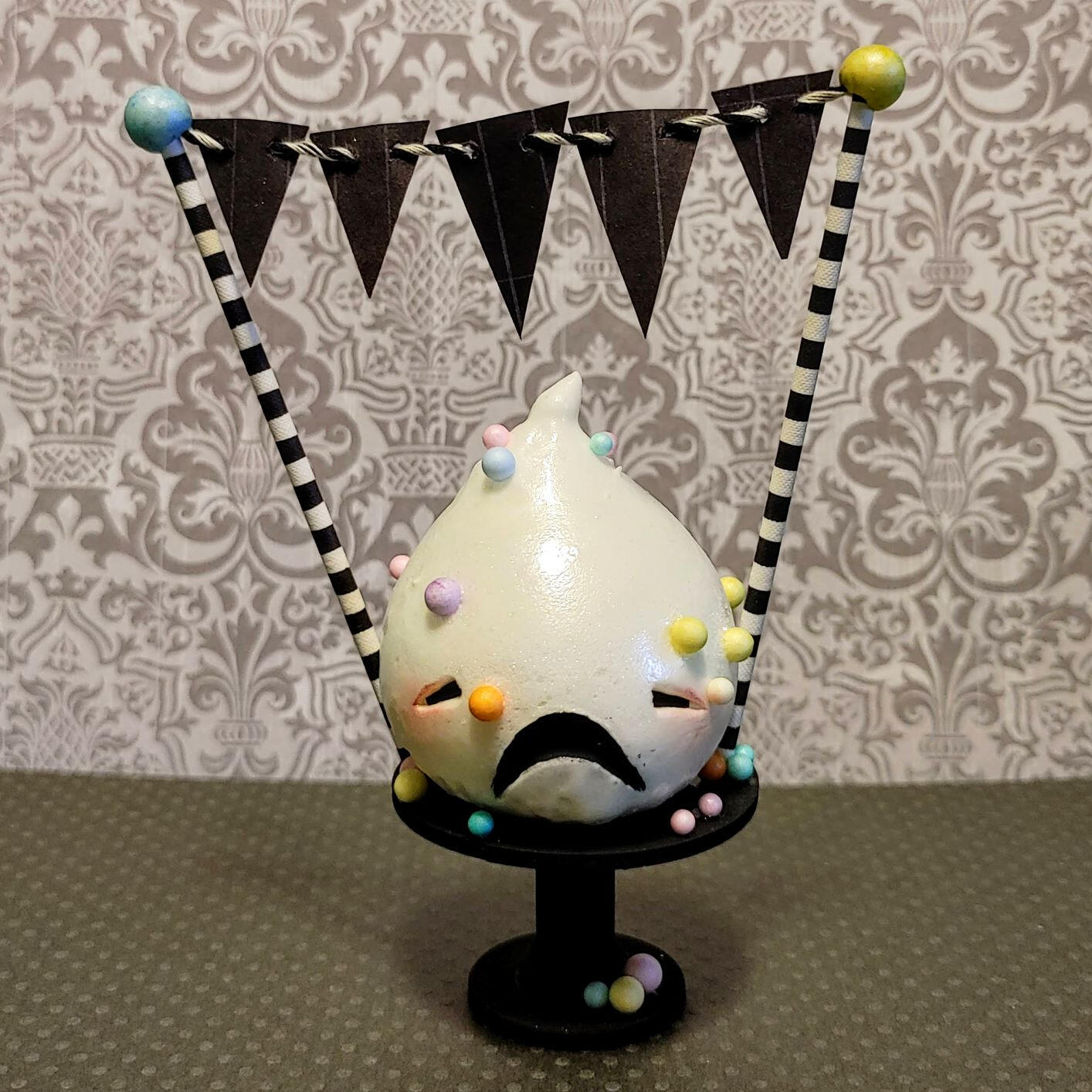 Small handmade sculpture of a distressed frosting creature covered in colorful sprinkles, displayed on a tiny cake pedestal.