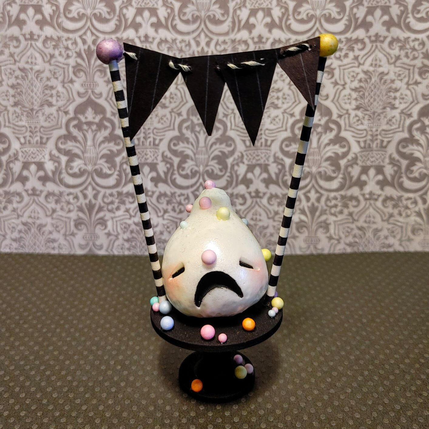 Small handmade sculpture of a distressed sprinkle-covered frosting creature on a tiny cake pedestal.