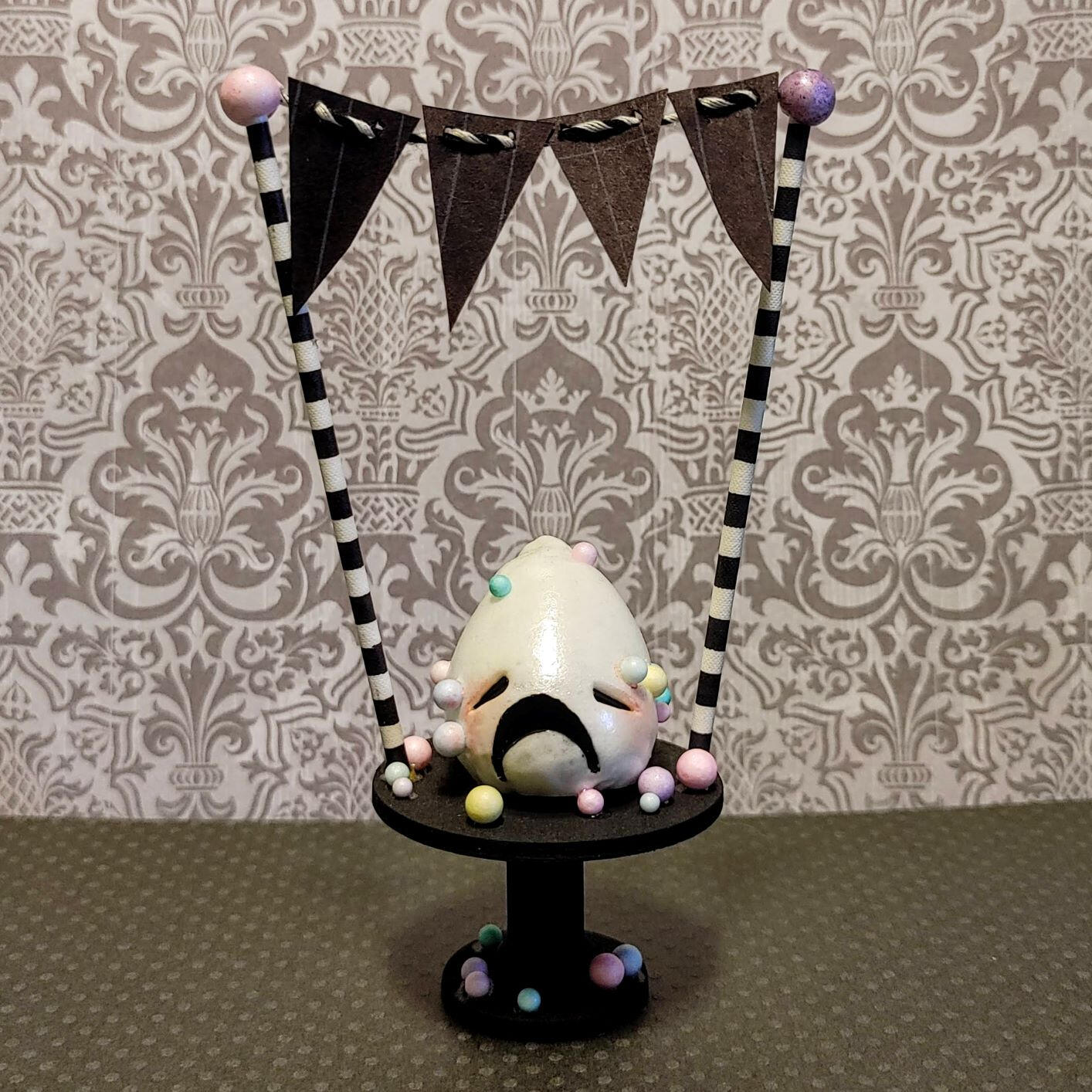 Small handmade sculpture of a distressed sprinkle-covered frosting creature perched on a tiny cake pedestal.