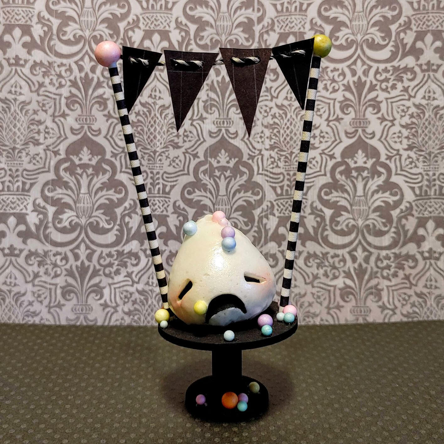Small handmade sculpture of a distressed blob of marshmallow frosting covered in colorful sprinkles and perched on a tiny cake pedestal with tiny black bunting flags topping striped decorative posts