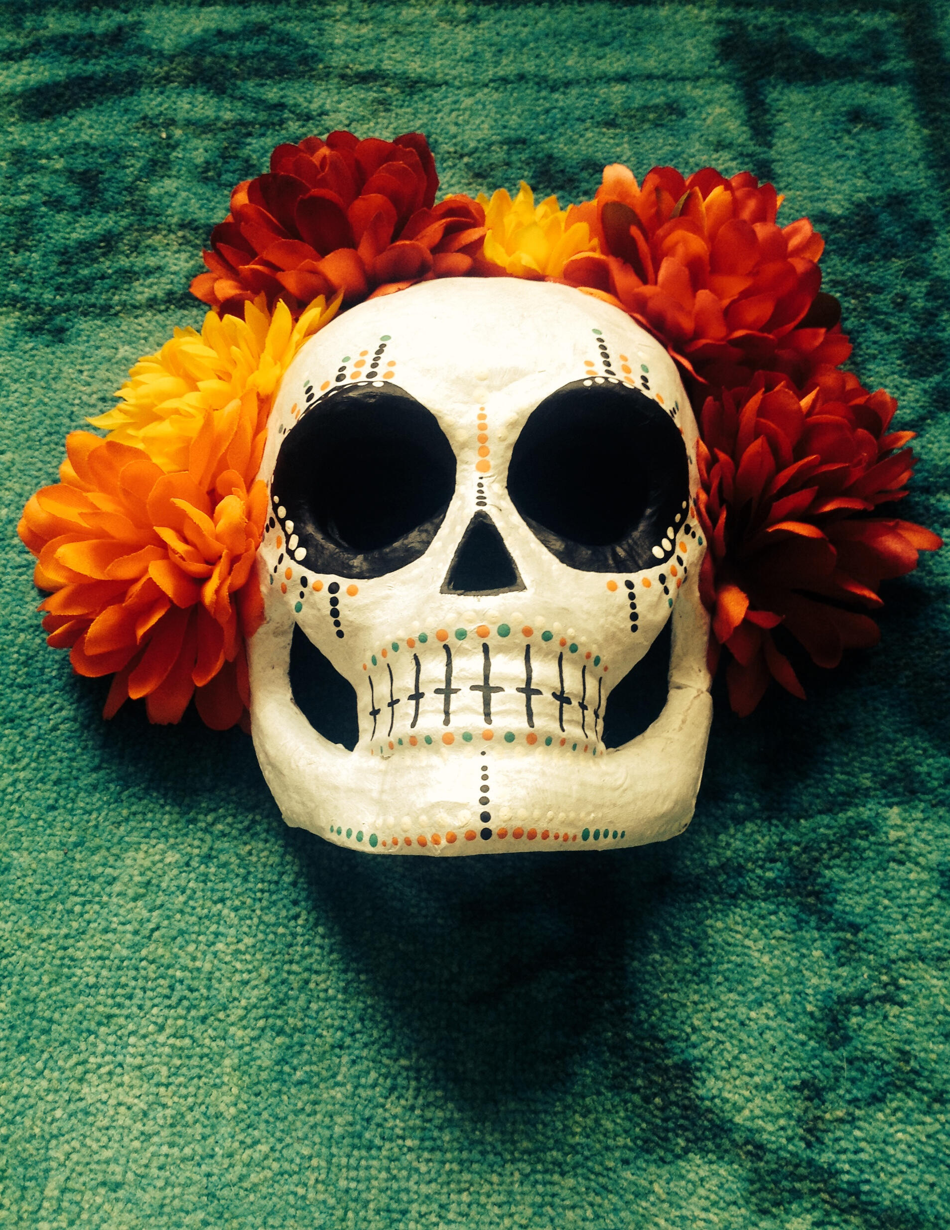 Paper-mâché skull mask decorated with colorful painted dots and crowned with large autumn-colored flowers.
