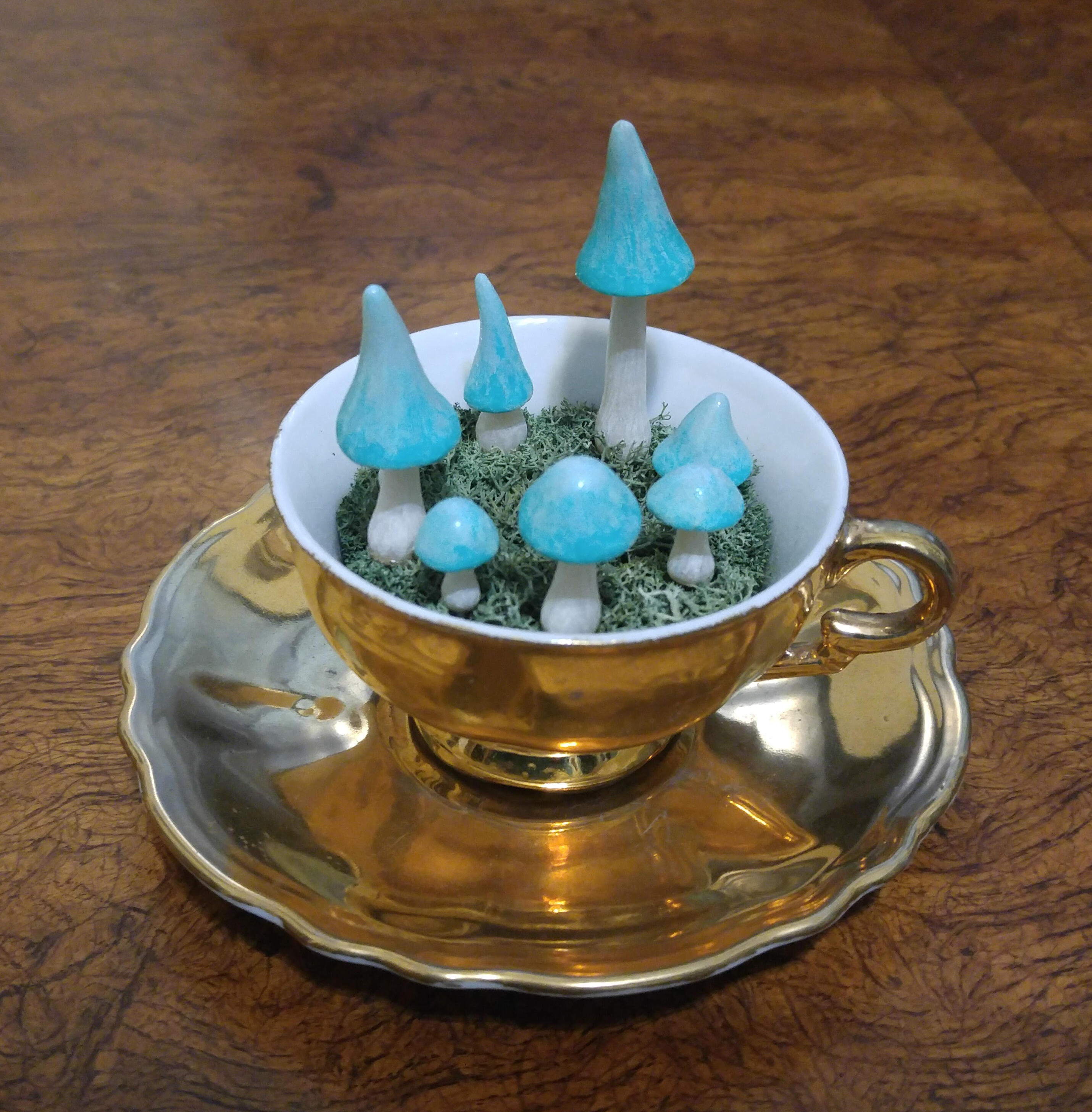 A tiny golden antique teacup filled with moss from which a circle of tiny turquoise mushrooms are sprouting in a ring