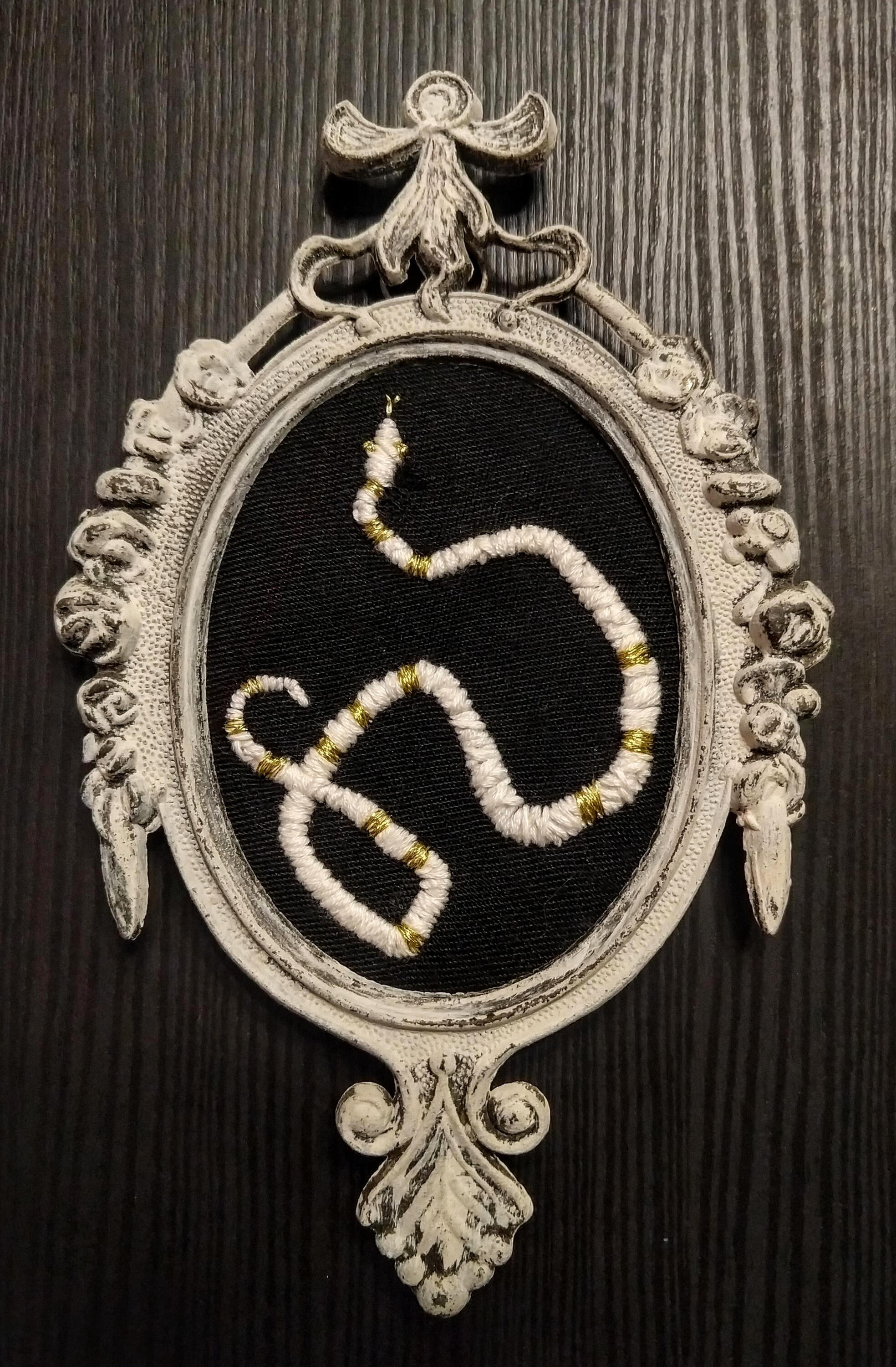 A white and gold embroidered snake on a black cloth background, framed in a cream colored brass antique victorian frame