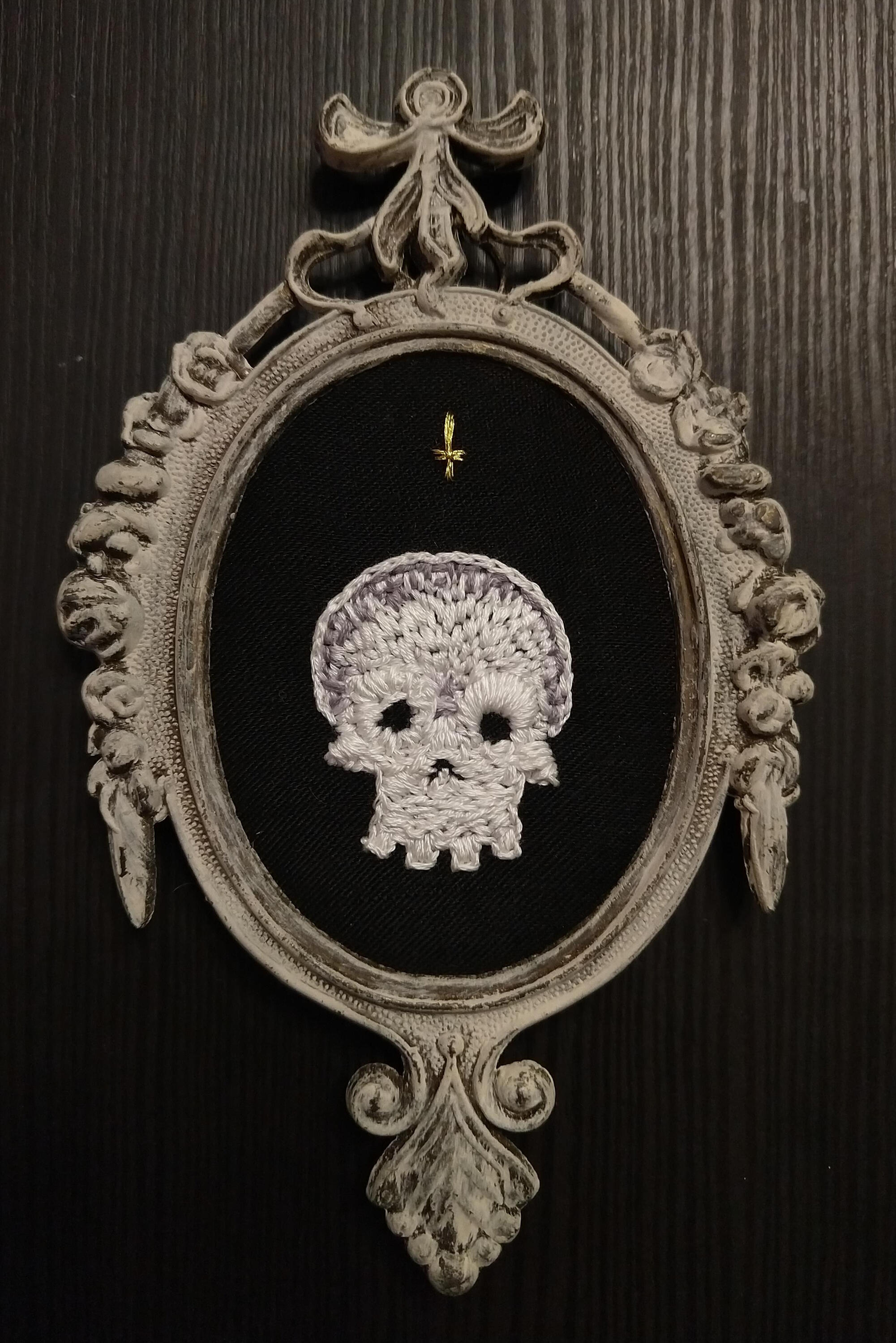 A white and gold embroidered skull on a black cloth background, framed in a cream colored brass antique victorian frame