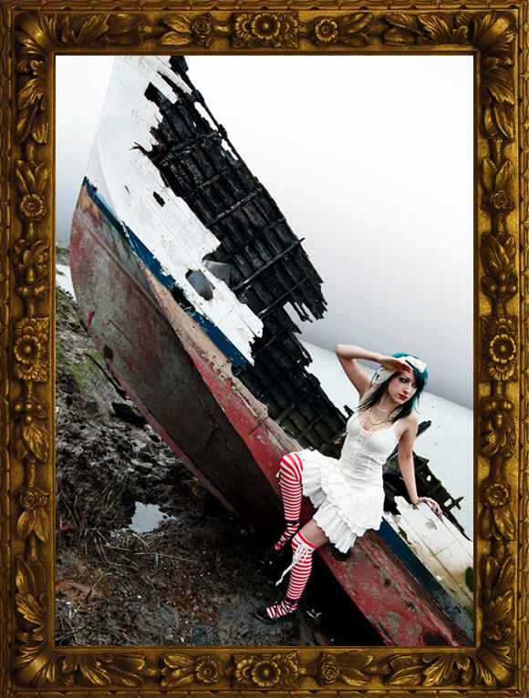 A mysterious individual with turquoise hair, wearing anachronistic cream colored bloomers and a button up dress with striped stockings and ballerina flats laced all around the ankles. A large lace bow is tied in their hair, and multiple strands of beaded