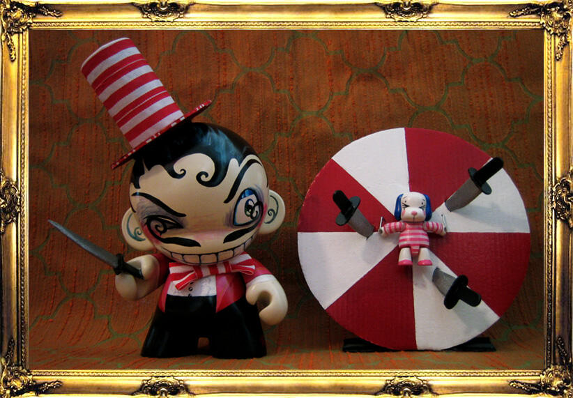 A Munny and Dunny, painted to be circus performers - a grinning knife thrower wearing a tall red and white striped hat holding a knife and squinting sideways at their assistant - a small blue eared dog wearing a matching outfit, tied to a spinning red and